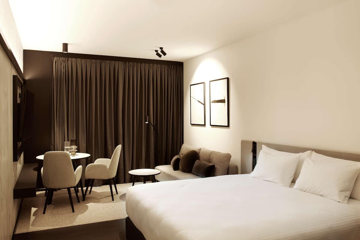 A modern hotel room with a large bed, a desk with chairs, a sofa, and two framed pictures on the wall.