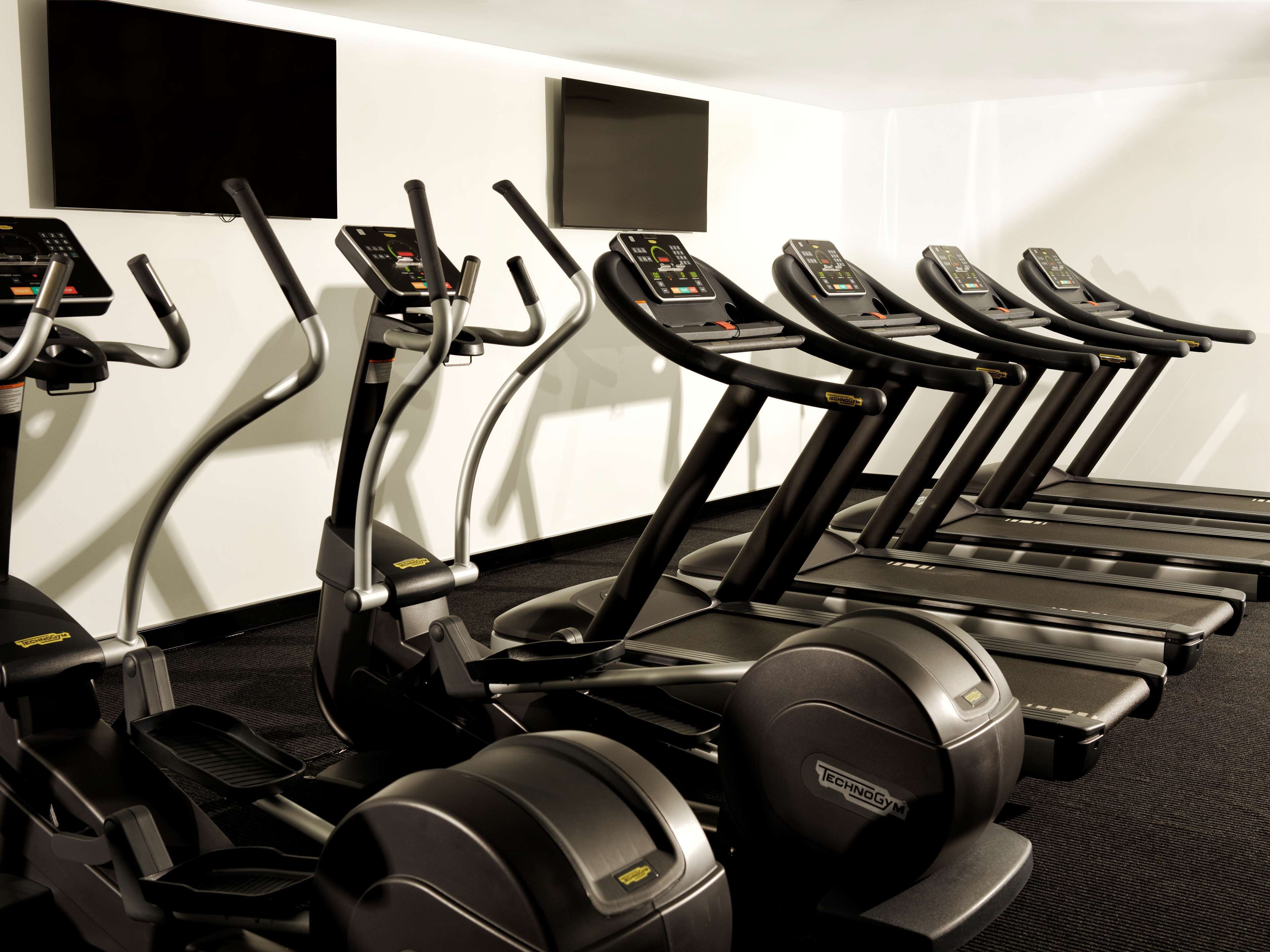 Une rangée de tapis de course dans une salle de sport, avec un téléviseur à écran plat fixé au mur.