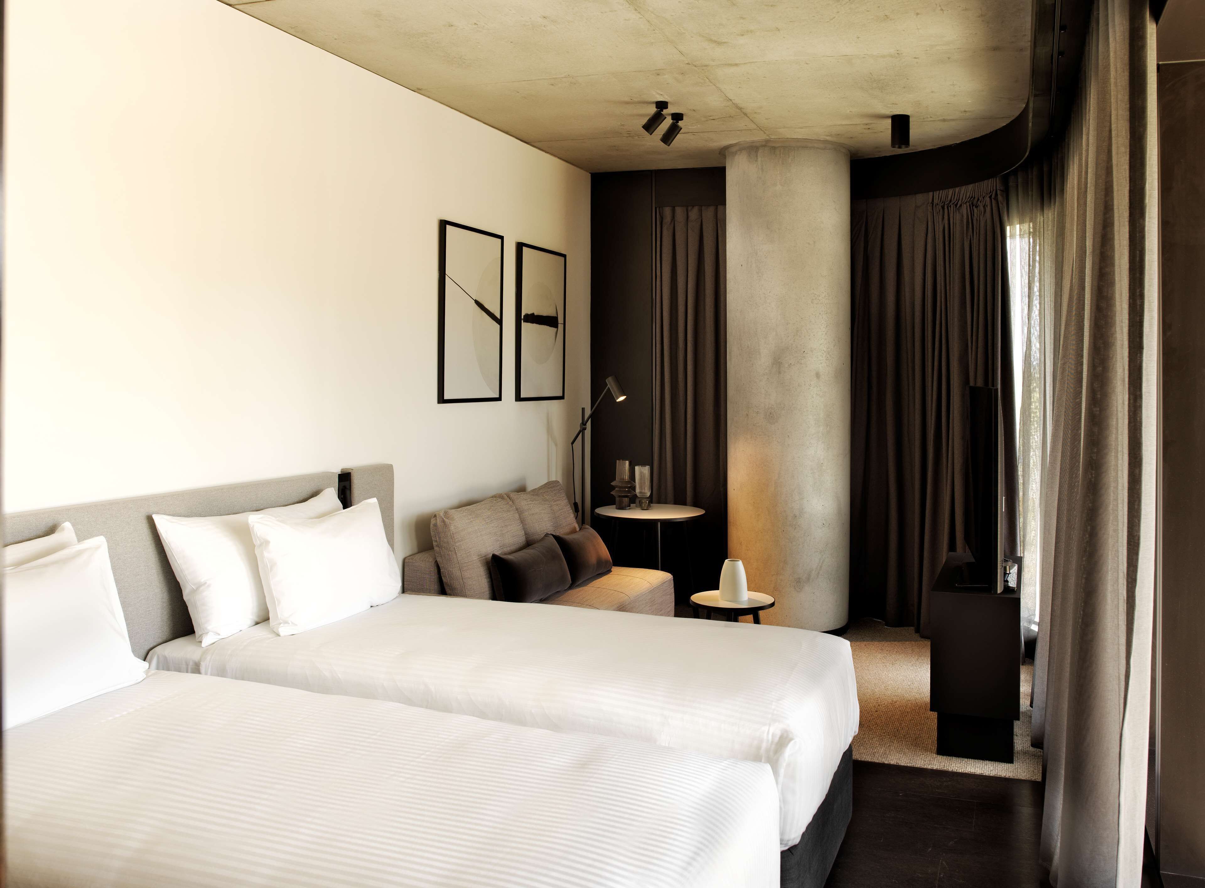 A hotel room with two beds, a small table, a lamp, a large pillar, and a view of the outdoors.