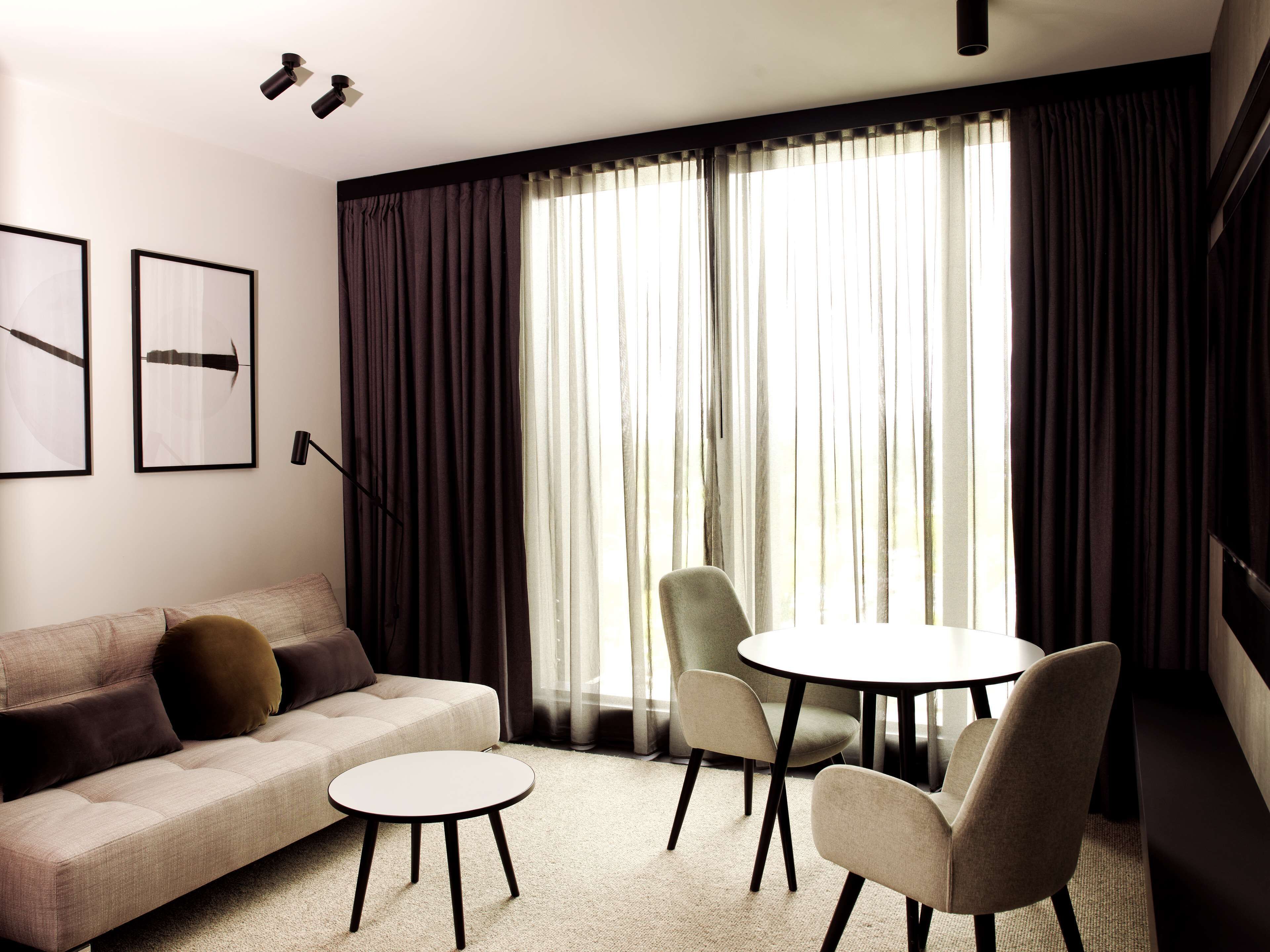 Ein modernes Wohnzimmer mit einer Couch, zwei Sesseln, einem kleinen runden Tisch und einem großen Fenster mit Vorhängen.