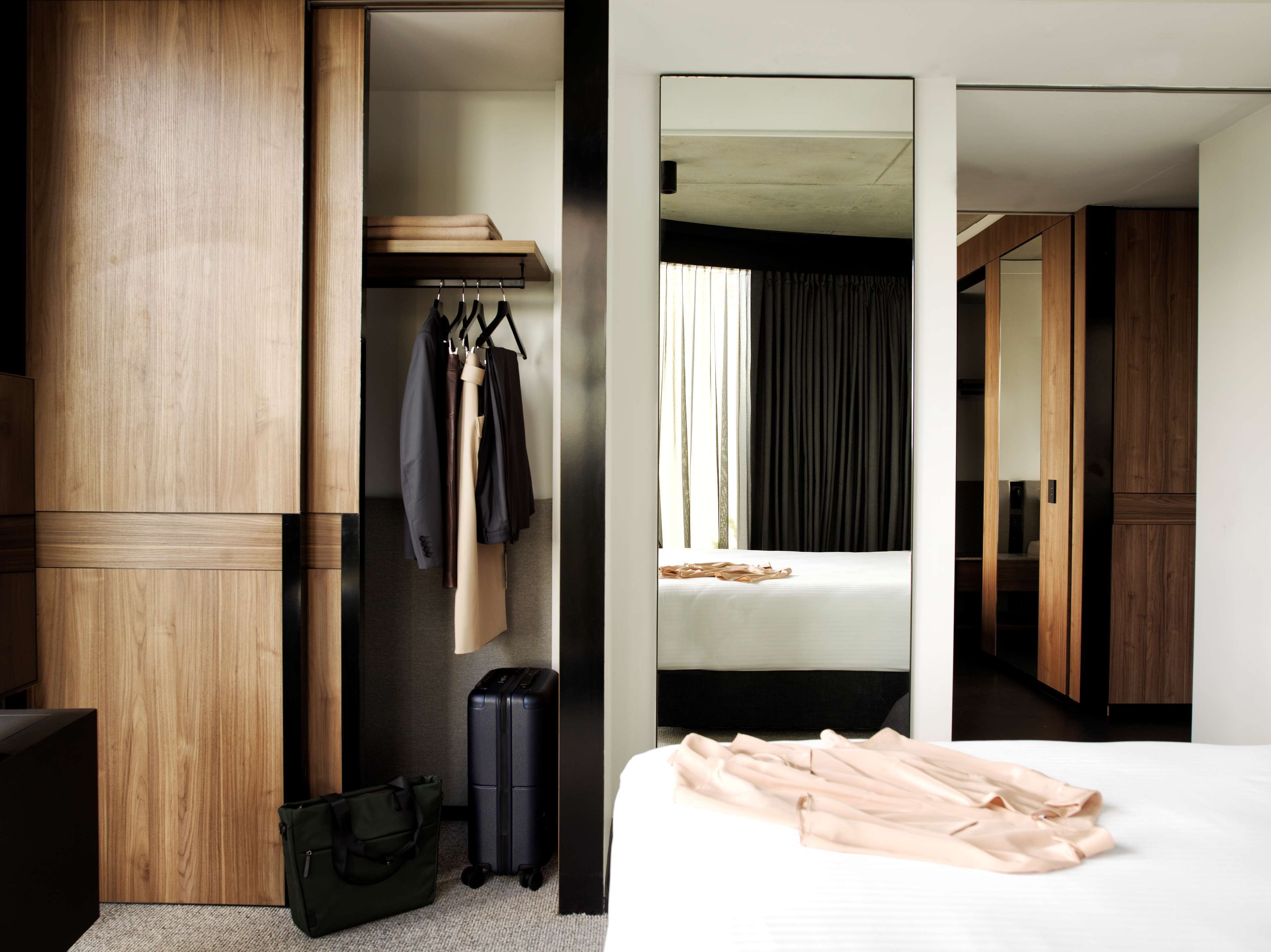 Ein Hotelzimmer mit einem Holzschrank, einem Bett mit weißer Matratze, einem Kleiderständer mit Kleidung, einem Koffer und einer Tasche auf dem Boden.