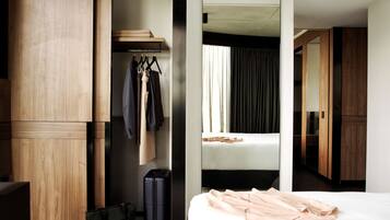 Ein Hotelzimmer mit einem Holzschrank, einem Bett mit weißer Matratze, einem Kleiderständer mit Kleidung, einem Koffer und einer Tasche auf dem Boden.