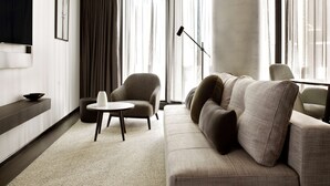 Un salon moderne avec un canapé, une petite table ronde, un téléviseur fixé au mur et de grandes fenêtres ornées de rideaux légers.