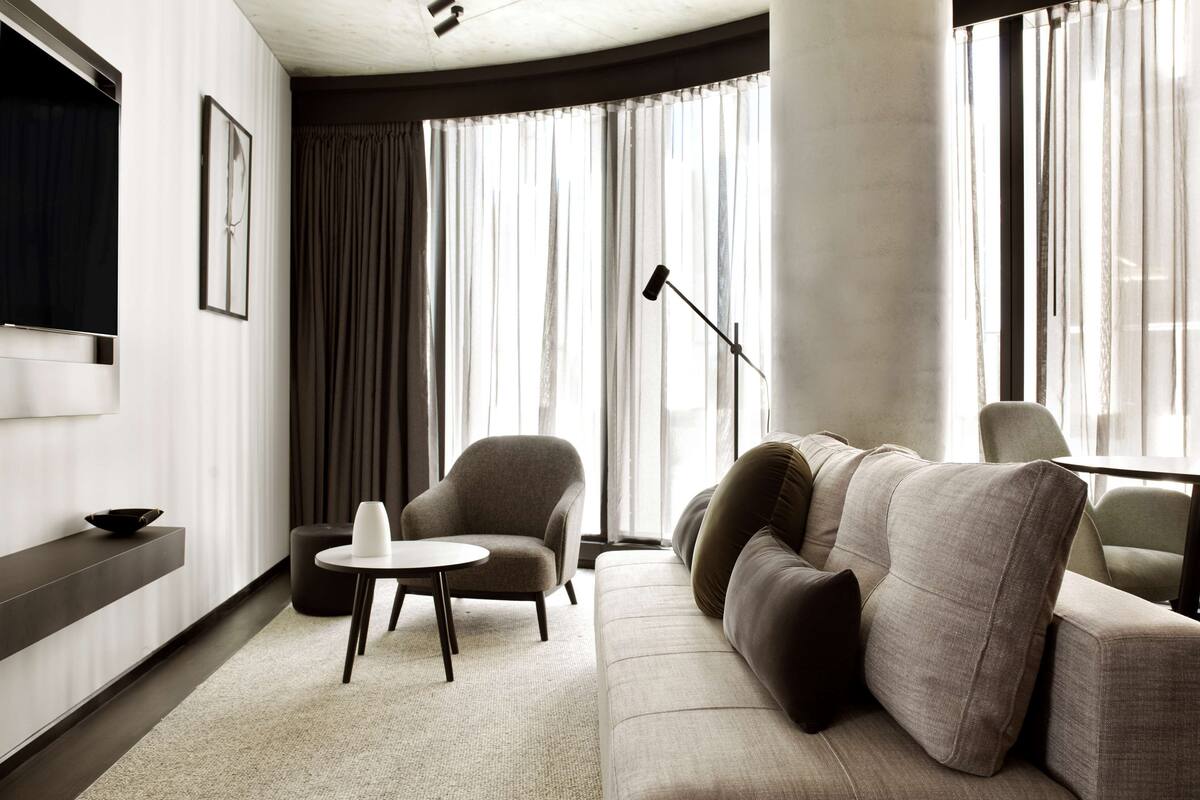 A modern living room with a sofa, a small round table, a TV mounted on the wall, and large windows with sheer curtains.