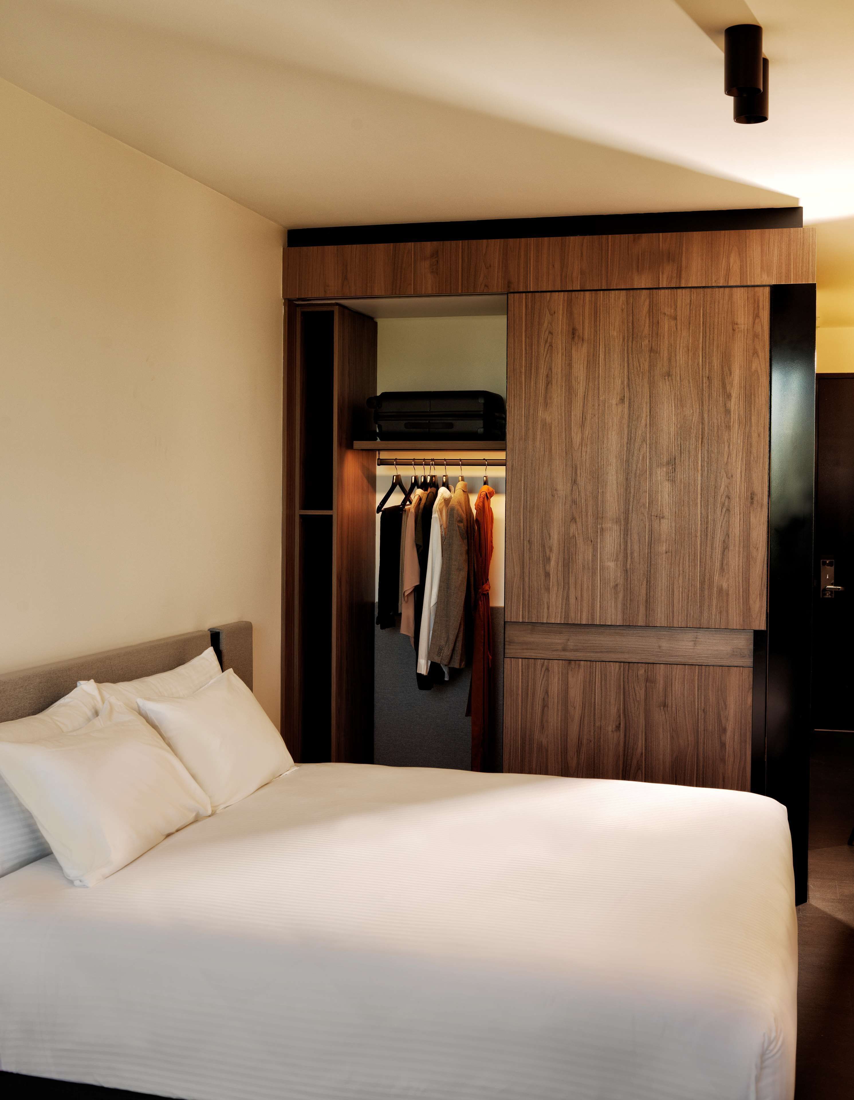 Quarto de hotel com uma cama grande, um guarda-roupa e um cabideiro com diversas peças de roupa.