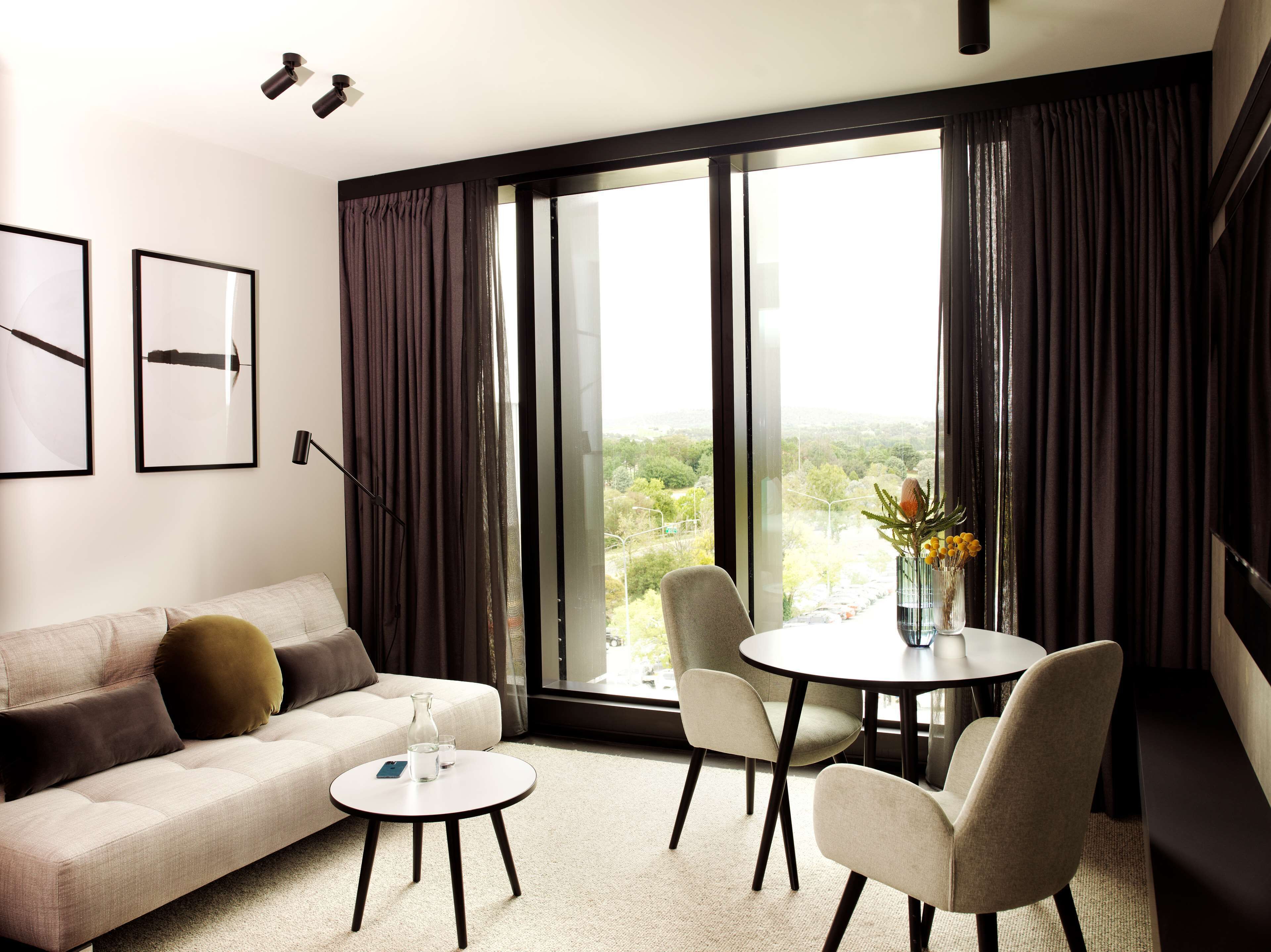 A modern hotel room with a sofa, a small round table, two chairs, a large window with curtains, and framed artwork on the wall.
