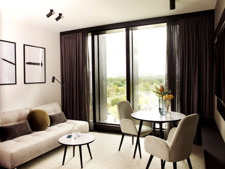 Moderni hotellihuone, jossa on sohva, pieni pyöreä pöytä, kaksi tuolia, suuri ikkuna verhoilla sekä seinällä tauluja.