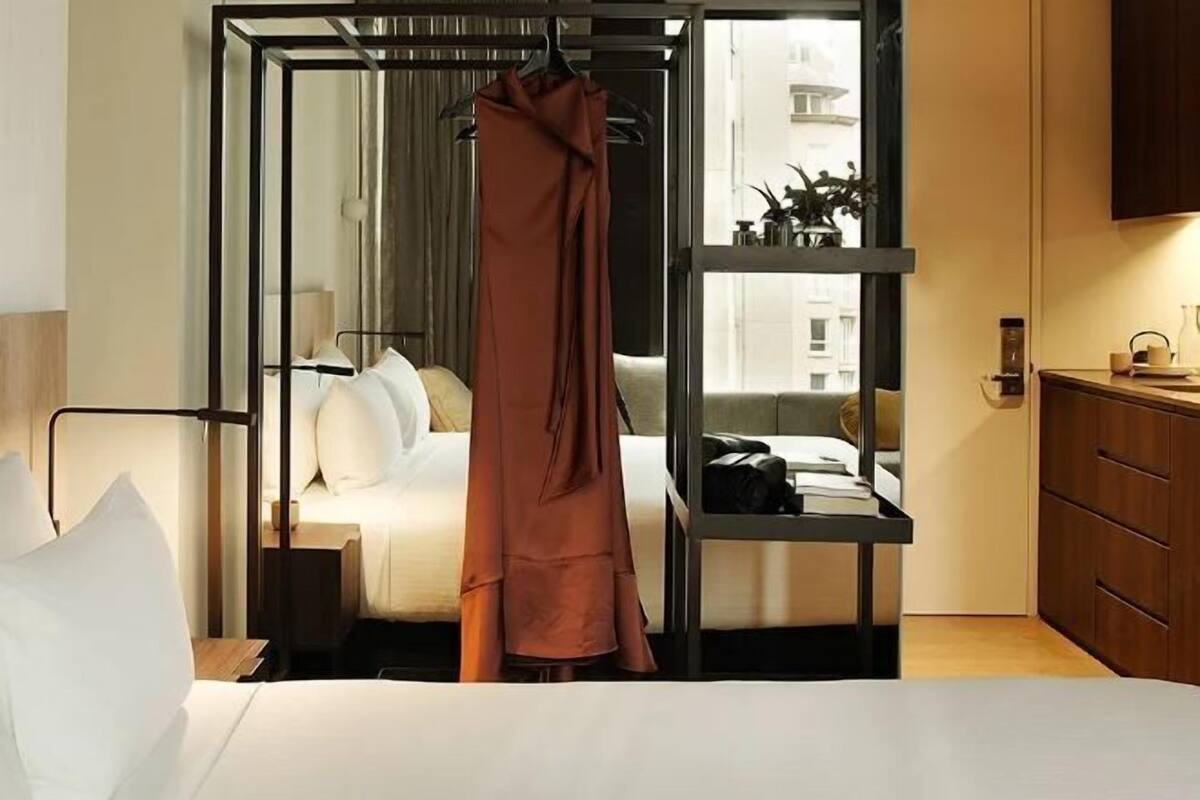 A modern hotel room with a bed, a hanging robe, a bedside table, and a view of the city through a window.