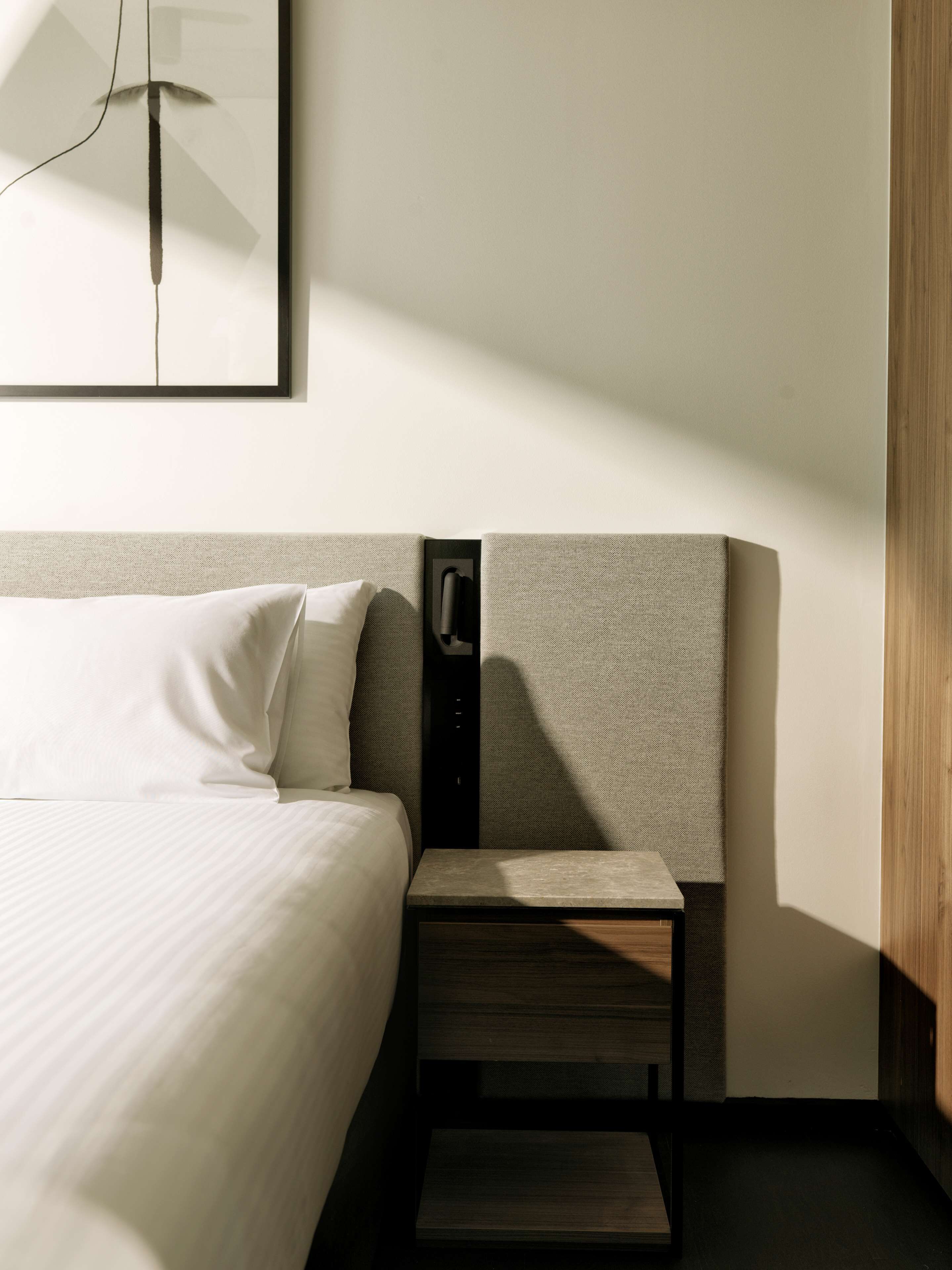 A modern hotel room with a neatly made bed, a small bedside table, and a wall-mounted lamp.