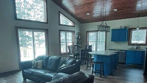 Un amplio salón con techo de madera, ventanas grandes, un sofá y una zona de cocina.