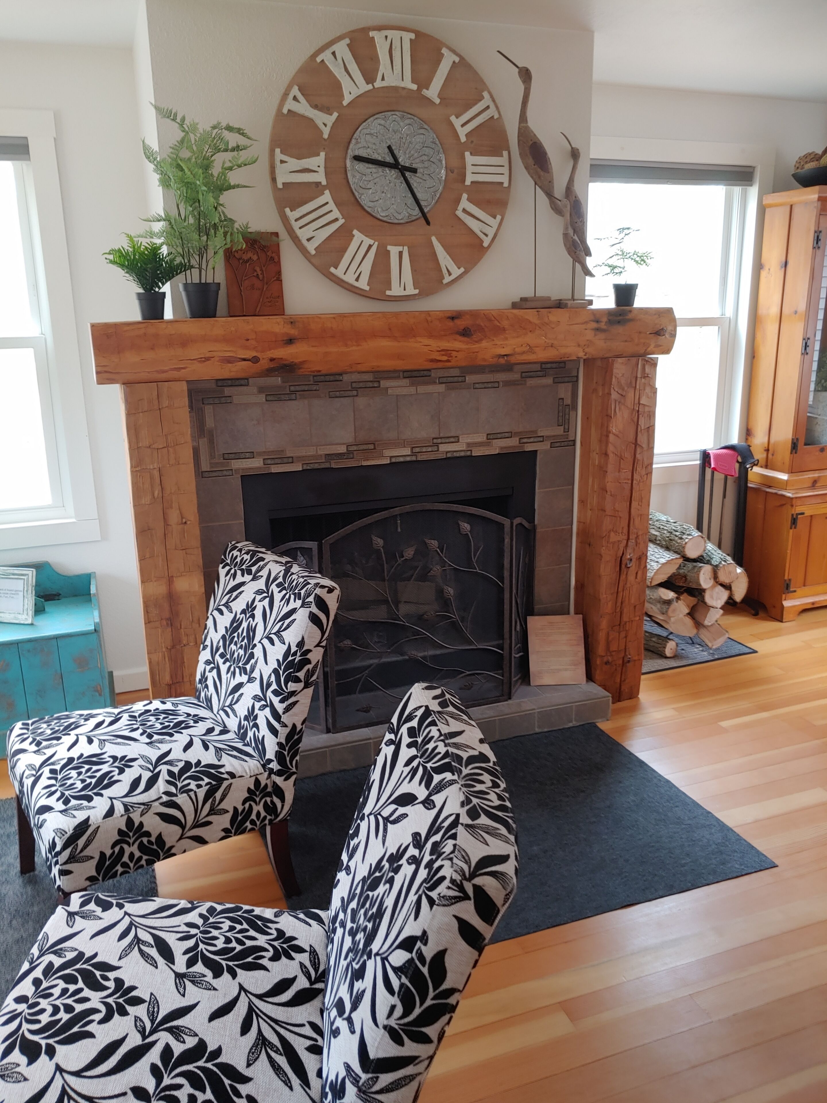 Un salon doté d’une cheminée, de deux fauteuils à motifs, d’une grande horloge murale et d’une cheminée en bois.