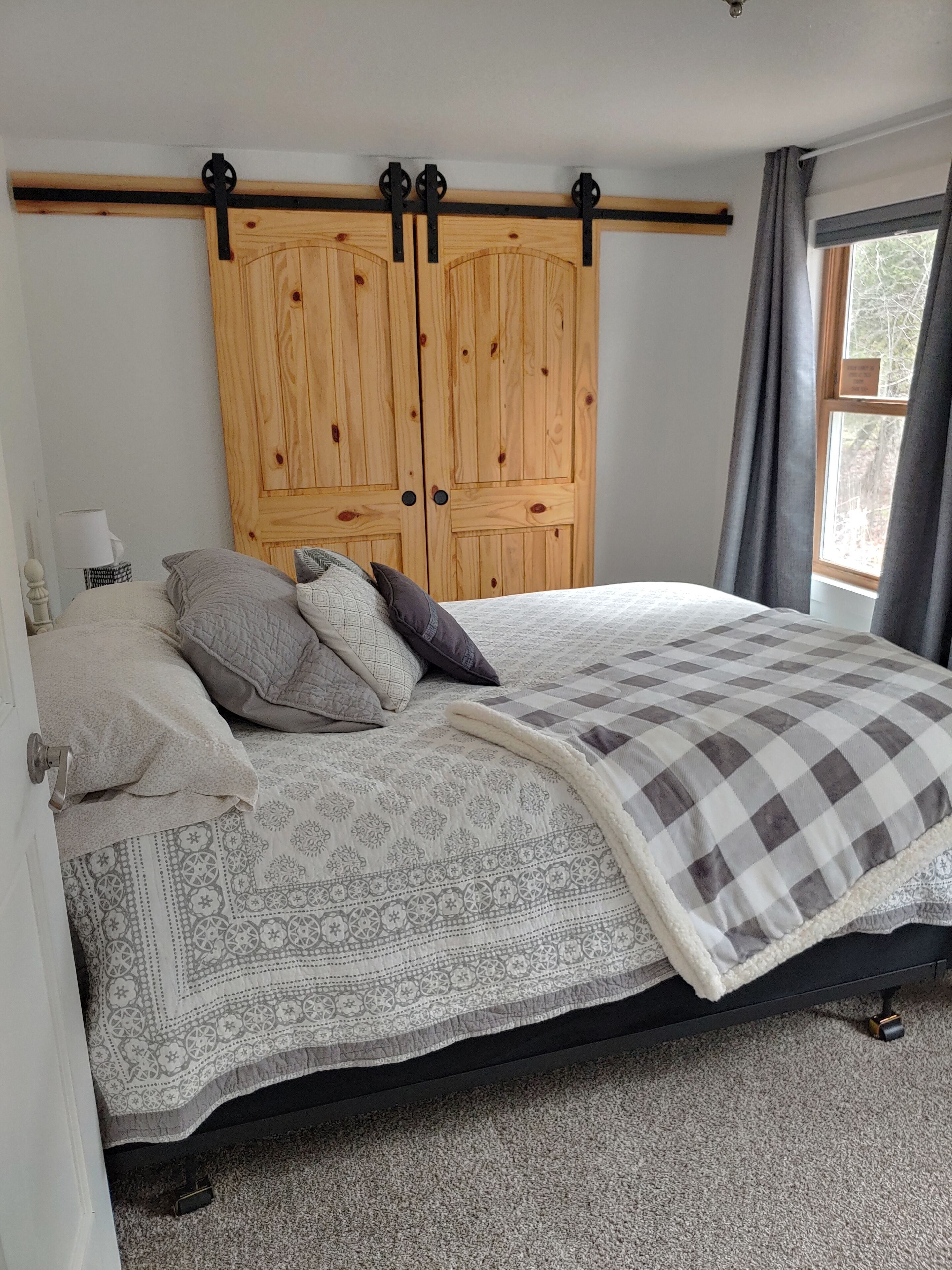 Ein Schlafzimmer mit einem Bett, Kissen, einer gemusterten Tagesdecke, einem Holzschrank und einem Fenster mit Vorhängen.
