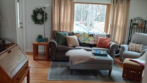 A living room with a sofa, armchairs, a coffee table, and a rug. There is a door with a wreath, a small table, and a window with curtains.