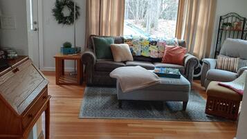Una sala de estar con sofá, sillones, una mesa de centro y una alfombra. Hay una puerta decorada con un adorno, una pequeña mesa y una ventana con cortinas.