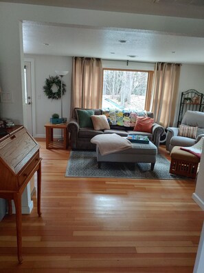 A living room with a sofa, armchairs, a coffee table, and a rug. There is a door with a wreath, a small table, and a window with curtains.