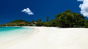 Ein Strand mit klarem türkisblauem Wasser, weißem Sand und einer kleinen Anhöhe mit Gebäuden im Hintergrund.