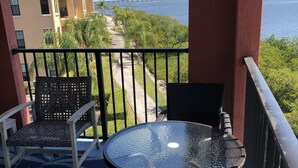Un balcon avec une table en verre et des chaises en osier, donnant sur une route côtière et un pont.
