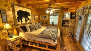 A log cabin bedroom with a wooden bed, a fireplace, a television, and a bear mural on the wall.