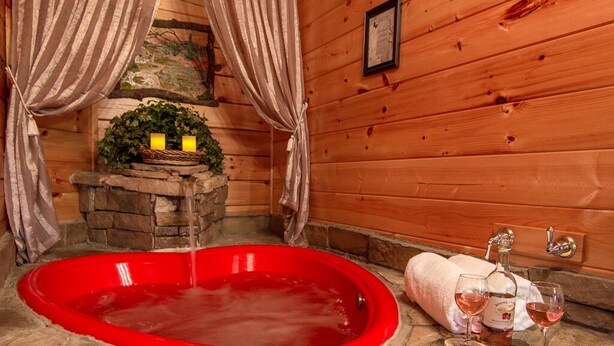 A wooden cabin interior with a red hot tub, a stone fireplace, and a bottle of wine on a wooden coaster.