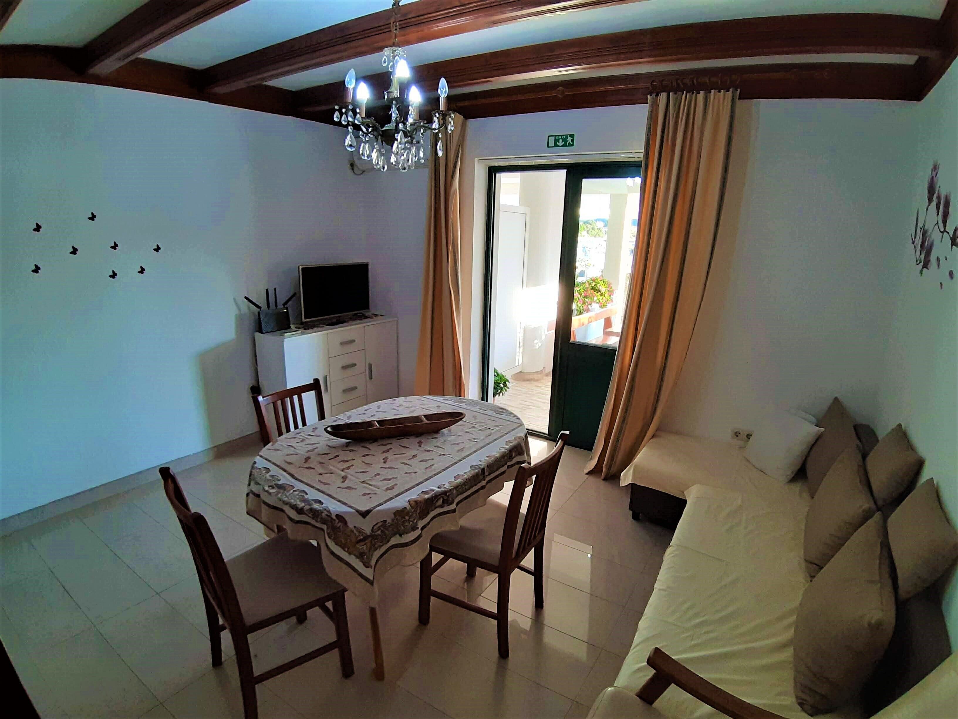 A dining area with a table, chairs, a chandelier, a television on a cabinet, and a sofa with cushions.