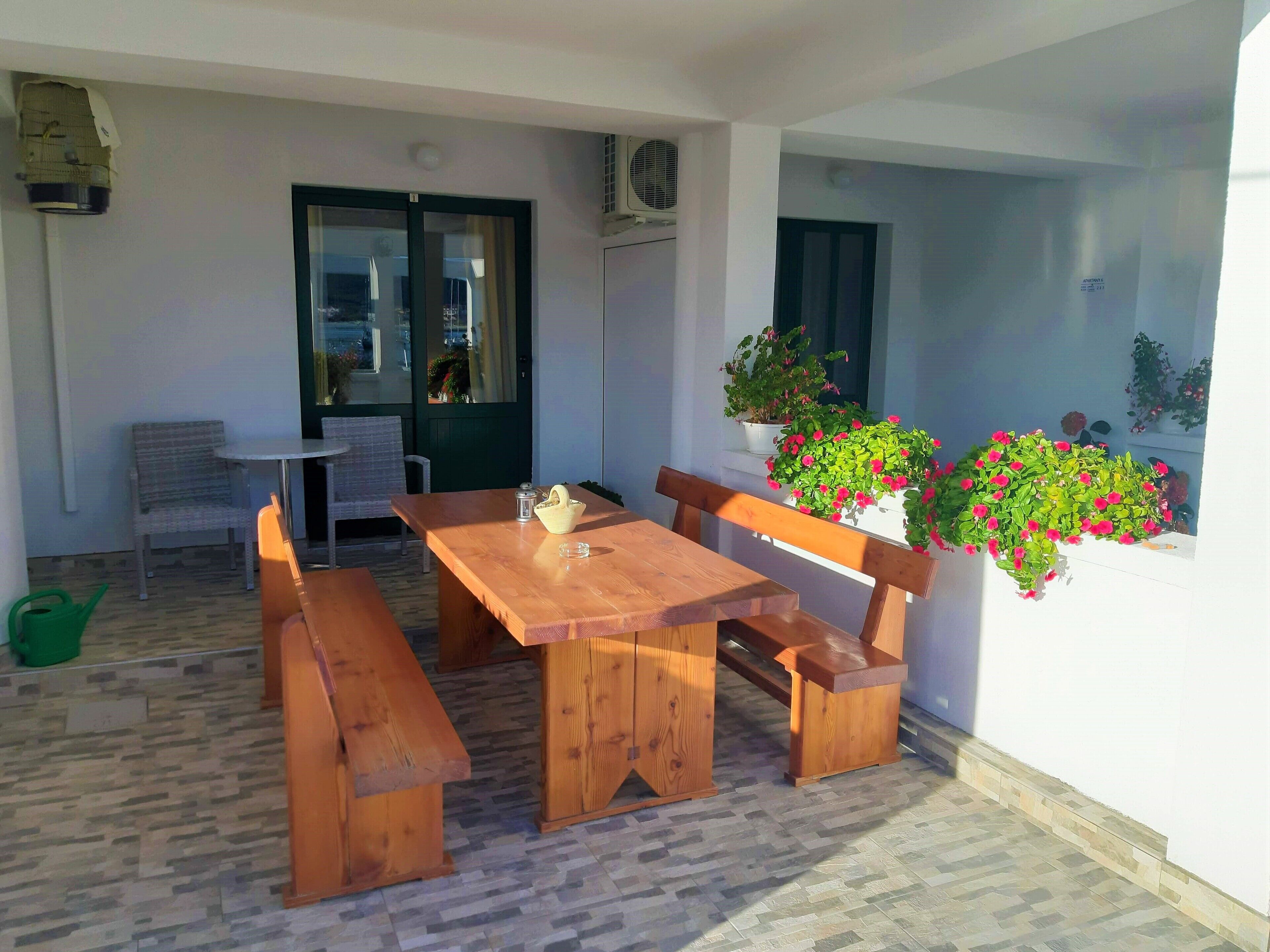 A wooden table with benches, a green watering can, and potted plants on a balcony.