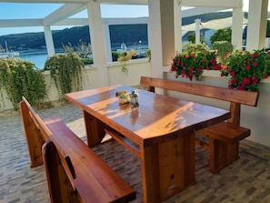 A wooden table and benches under a pergola with a view of a lake and boats. - Room in Apartment - Apartments and Rooms With a Swimming Pool Kampor - rab (Rab)