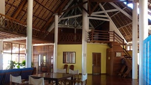 A spacious indoor area with a high ceiling, wooden beams, and a thatched roof visible through the windows. There is a seating area with tables and chairs, and a staircase leading to an upper level. - Spend all day on the Beach and Enjoy the Amenities Offerd (Nosy Be)