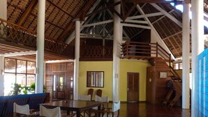 A spacious indoor area with a high ceiling, wooden beams, and a thatched roof visible through the windows. There is a seating area with tables and chairs, and a staircase leading to an upper level. - Nosy be, a Wonderful Location to Have a Relaxing Vacation (Nosy Be)