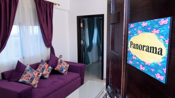 A living room with a purple sofa, colorful pillows, a glass coffee table, and a door with a 'Panorama' sign. - Panorama Apartment up to 3 Persons - Feel Home Away From Home (Dahab)