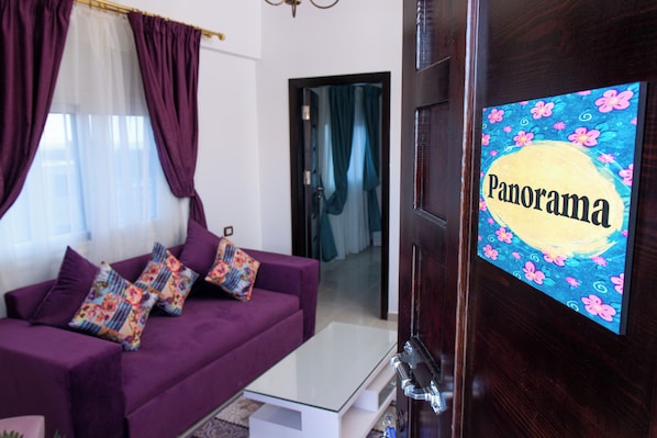 A living room with a purple sofa, colorful pillows, a glass coffee table, and a door with a 'Panorama' sign. - Panorama Apartment up to 3 Persons - Feel Home Away From Home (Dahab)