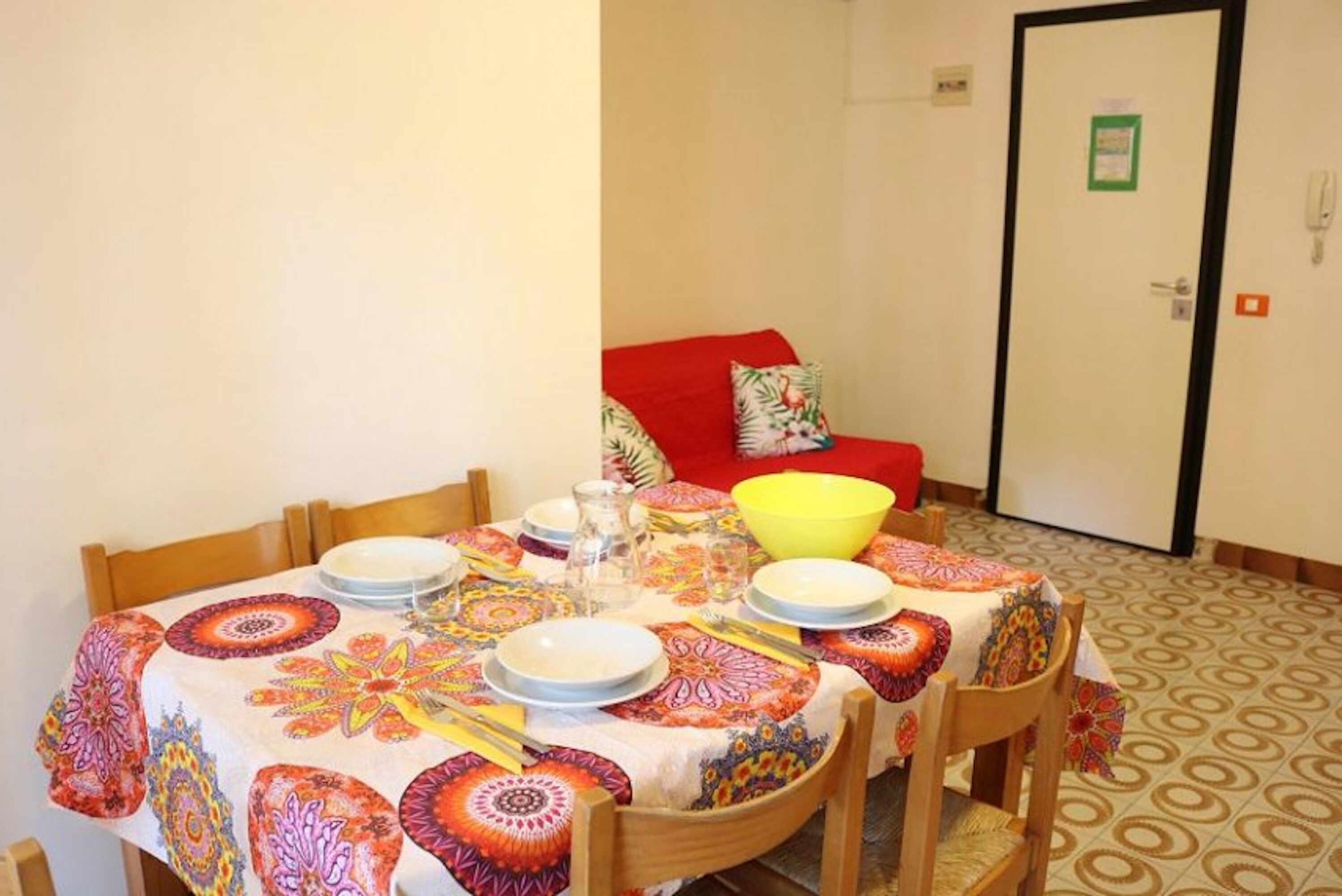 Un coin repas avec une table dressée pour quatre personnes, des chaises, un canapé rouge et une porte avec une enseigne verte.