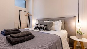 A neatly made bed with a grey and white duvet, a bedside table with a plant, and a hanging light fixture. - 2br Suite White gea With Private Pool No01 (Skiathos)