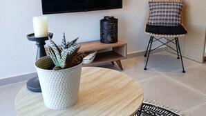 A modern living room with a flat-screen TV mounted on the wall, a round wooden table with a patterned rug, a wicker chair, and a small side table with a candle and a potted plant. - 2br Suite White gea With Private Pool No01 (Skiathos)