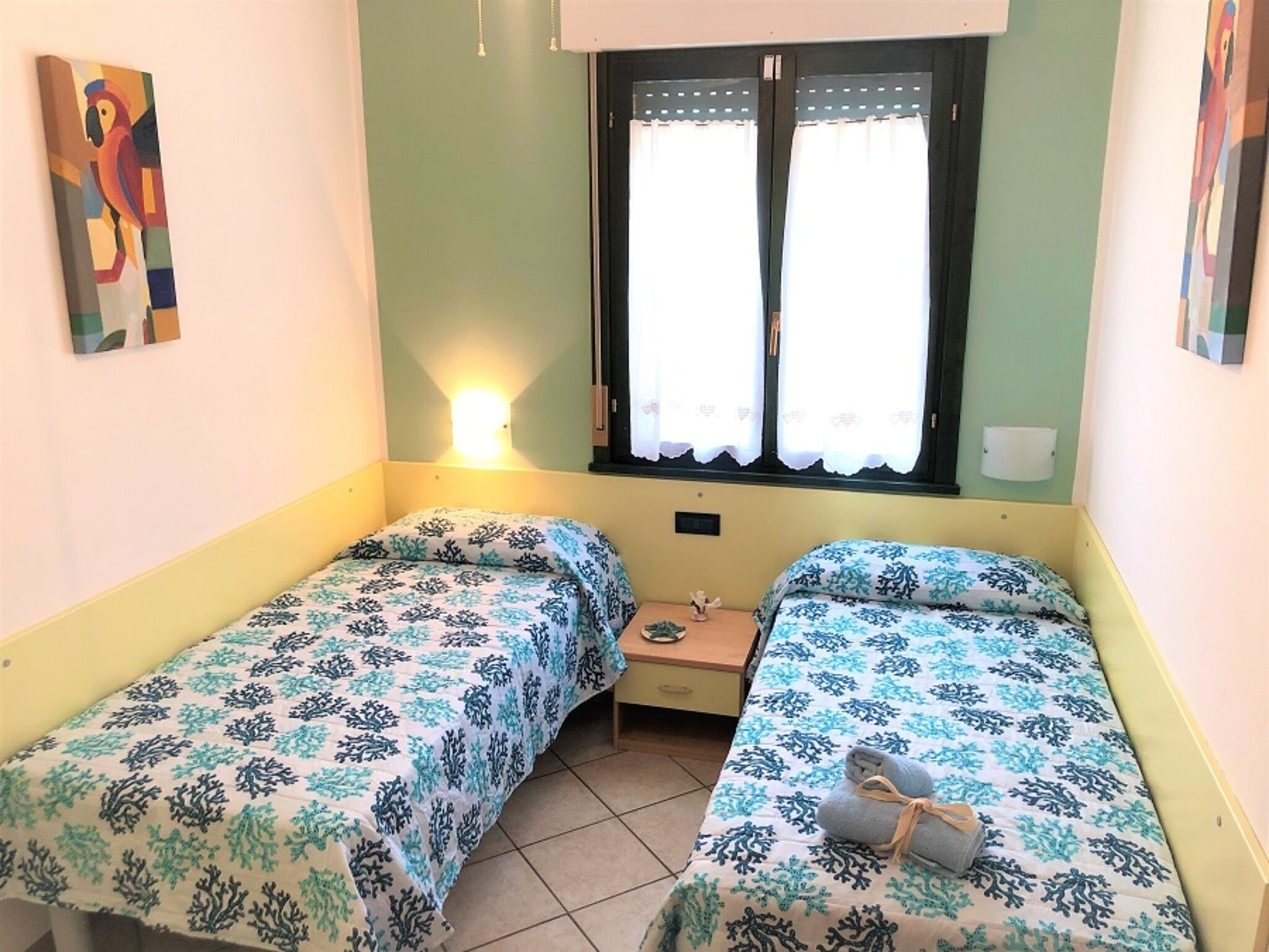 A small, single-room accommodation with two single beds, a nightstand, a window with curtains, and a wall-mounted light.