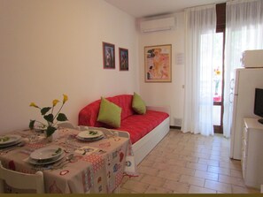 A room with a dining table set for two, a red sofa with green cushions, a television, and a painting on the wall.