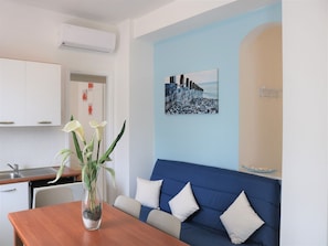 Un salon moderne avec un canapé bleu, une table à manger en bois, un vase rempli de fleurs et un tableau représentant un paysage urbain accroché au mur.