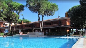 A swimming pool with a tiled deck, surrounded by trees and a building with balconies. - Flat in a Poolside Residence-beahost (San Michele al Tagliamento)