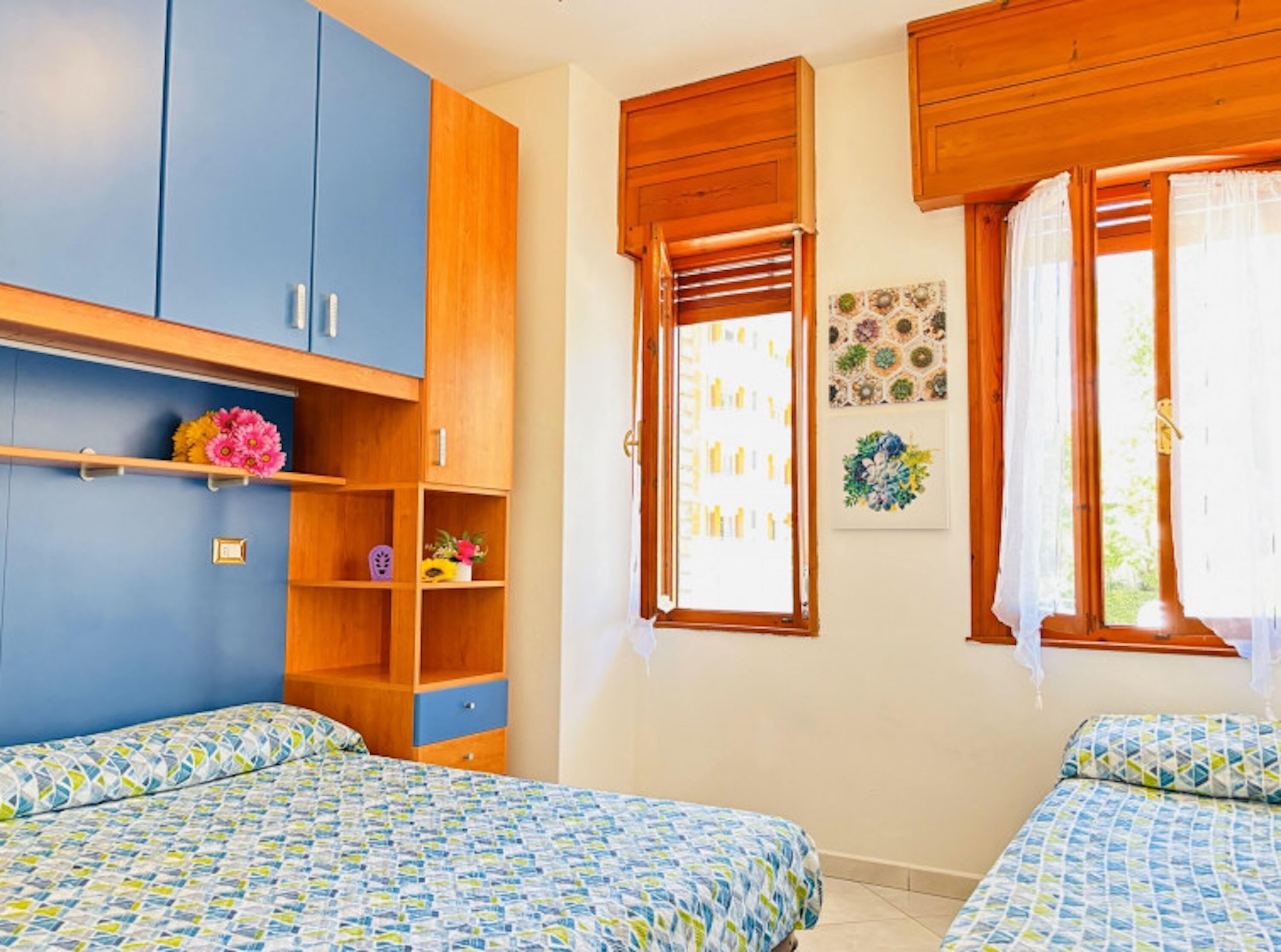 A bedroom with two beds, wooden cabinets, a shelf with flowers, and a window with curtains.