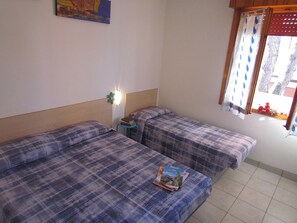A room with two beds, a small table with books, a wall-mounted light, and a window with curtains. - Apartment in Exclusive Holiday Complex (San Michele al Tagliamento)