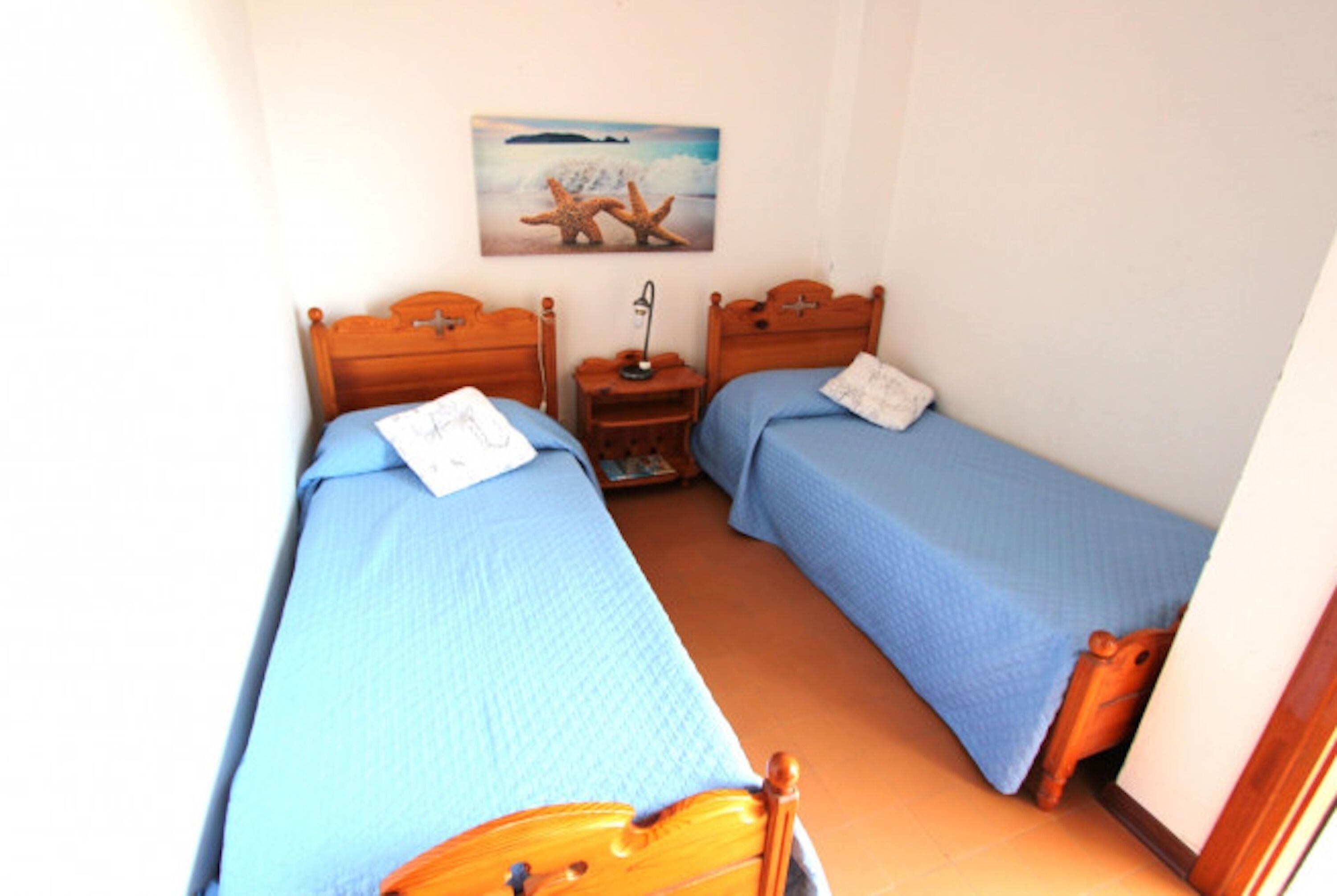 A small bedroom with two single beds, a nightstand, a lamp, and a painting on the wall.