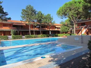 Une piscine dont les bords sont recouverts de carreaux, entourée d’une terrasse également carrelée. On aperçoit des arbres et un bâtiment avec un toit en tuiles à l’arrière-plan.