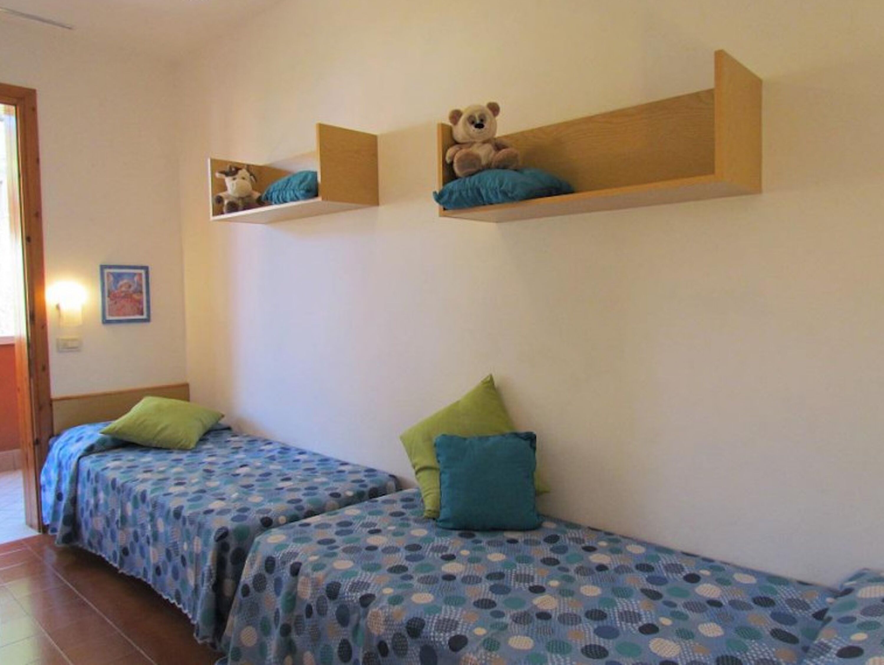 Une chambre avec deux lits, chacun recouvert d’une couverture à pois bleus et agrémenté d’oreillers verts. Au-dessus des lits, on trouve des étagères en bois sur lesquelles sont posés des ours en peluche.