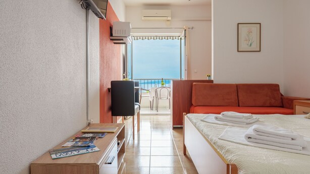 Una camera d'albergo con un letto, una TV, una scrivania con riviste, un balcone con vista e una finestra con condizionatore.