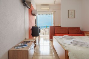 A hotel room with a bed, a TV, a desk with magazines, a balcony with a view, and a window with an air conditioner. - Sea View Apartment Studio for 2-3 People - Close to Commodities (Baska Voda)