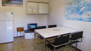 A compact kitchen with a white refrigerator, a small sink, and a dining table with four chairs. A television is mounted on the wall, and there is a painting of a beach scene above the refrigerator. - Lovely Flat With Garden and Shared Pool (San Michele al Tagliamento)