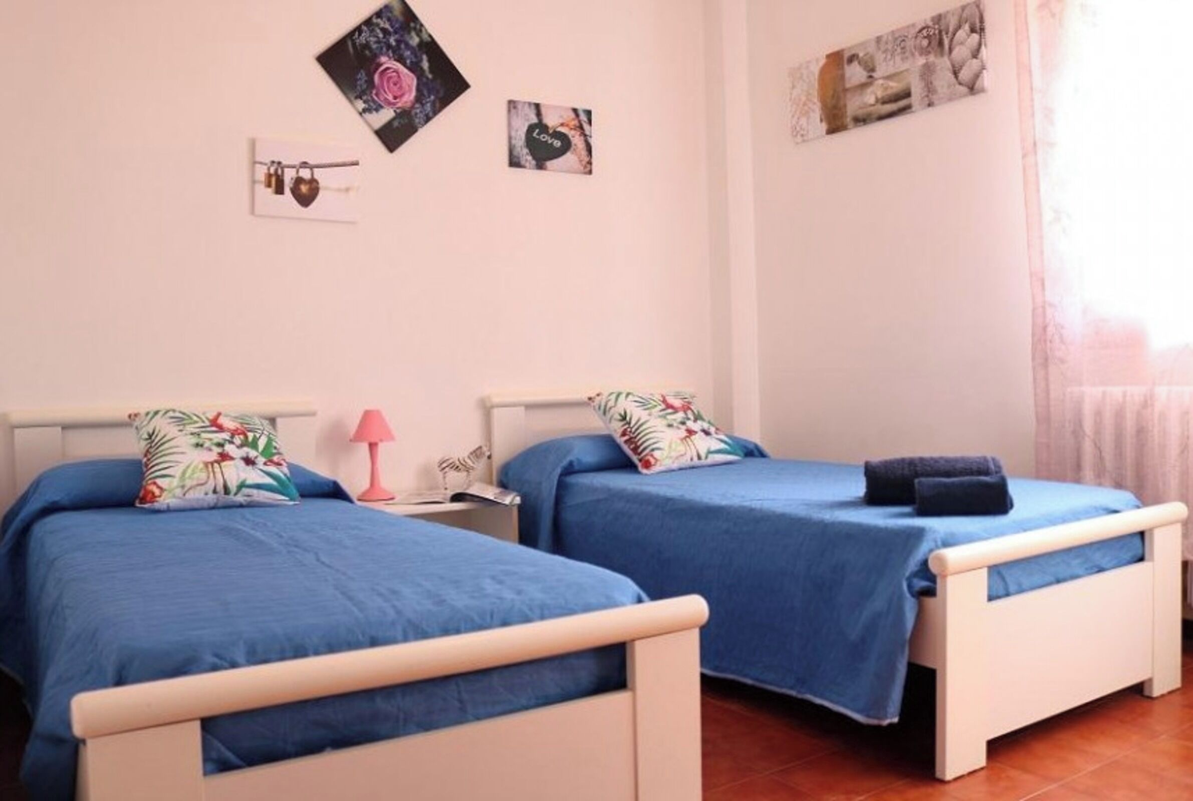A room with two single beds, each with a blue bedspread and floral pillows. There is a small table with a pink lamp between the beds. The walls are adorned with framed pictures, and the room has a tiled floor.