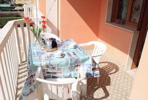 A balcony with a table set for two, white chairs, a tablecloth with a nautical theme, and a vase with red flowers.