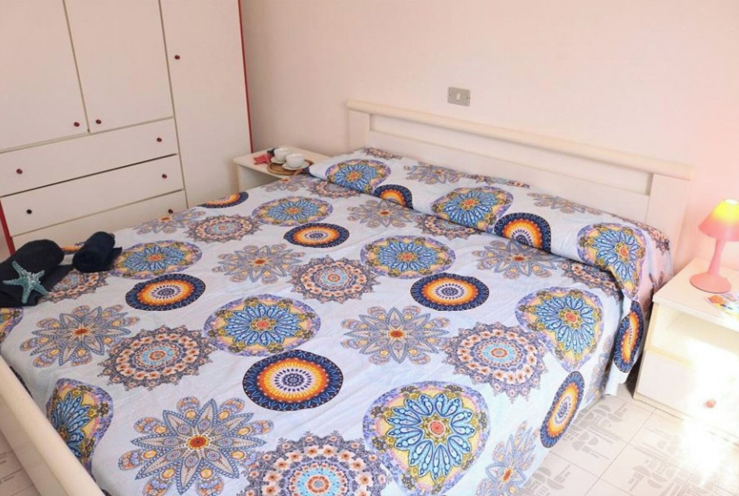 A bedroom with a bed covered in patterned bedding, a bedside table with a lamp, and a wardrobe.