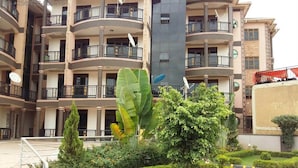 Un edificio residenziale a più piani con balconi, antenne paraboliche e un giardino curato con siepi e un albero al centro.