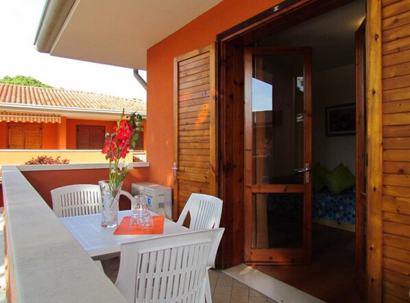 An outdoor dining area with a table and chairs, a vase with flowers, and a sliding glass door leading to a room with a bed and pillows.